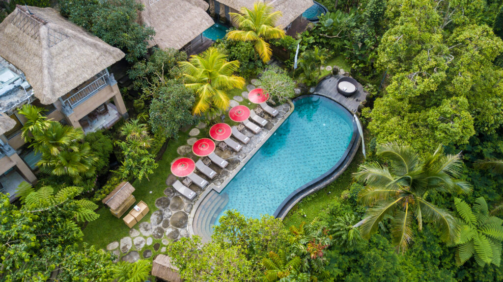 Aerial view of a private resort swimming pool surrounded by tropical palm trees, lounge chairs, and lush garden landscape in Bali.
