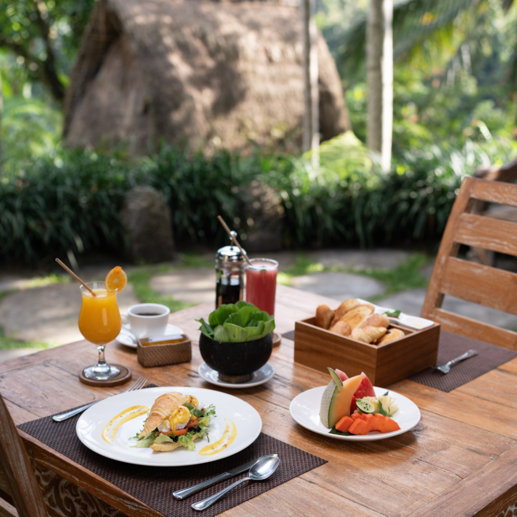Breakfast setup with fresh juice, coffee, and pastries served on a wooden table in a lush tropical garden setting in Bali.